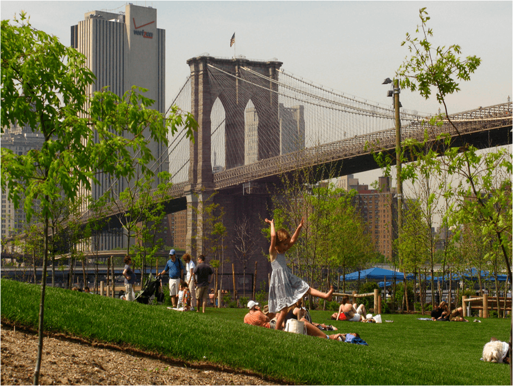 Child in foreground playing on grass with people in background laying down. Brooklyn Bridge in background with tall buildings.